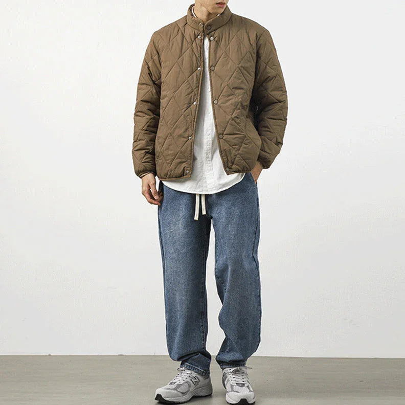 A person stands against a plain white background wearing a brown lightweight insulating padded jacket with a diamond-stitched pattern, a white shirt, loose blue jeans, and gray sneakers. Their hands are in their pockets and their face is not fully visible.