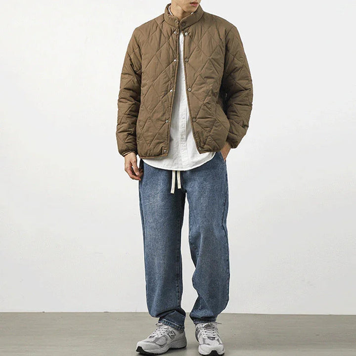 A person stands against a plain white background wearing a brown lightweight insulating padded jacket with a diamond-stitched pattern, a white shirt, loose blue jeans, and gray sneakers. Their hands are in their pockets and their face is not fully visible.