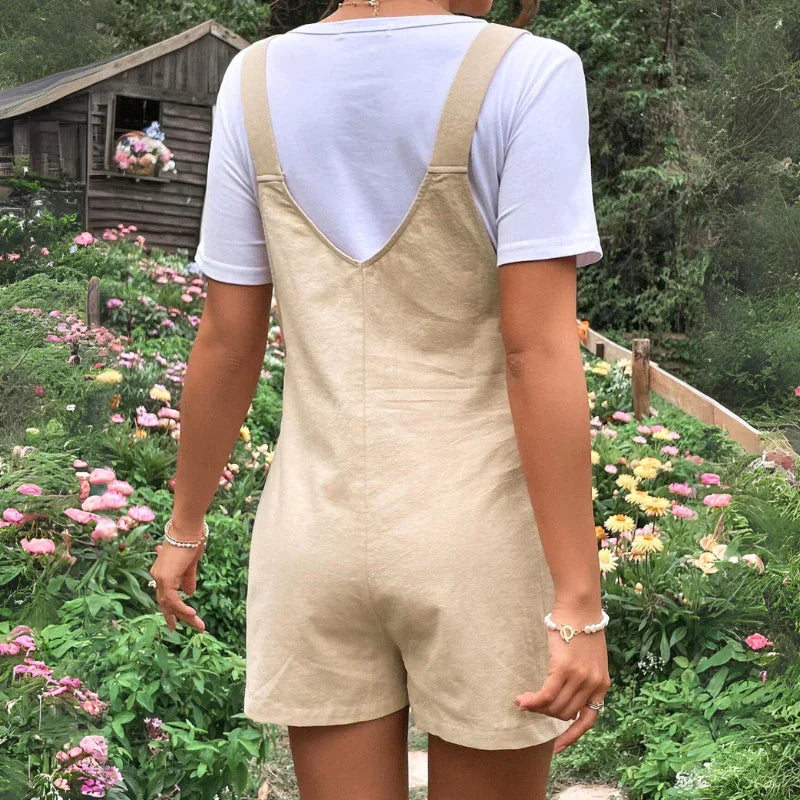 A person in comfortable maternity overalls and a white t-shirt stands in a flower garden, facing away from the camera. A wooden shed decorated with blooms is visible in the background.