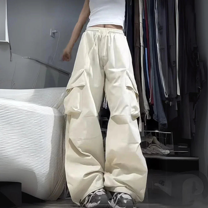 A person wearing a white sleeveless top and oversized beige cargo pants with cargo pockets stands in a bedroom, clothing hanging in an open closet behind them. Only the lower half of their face is visible.
