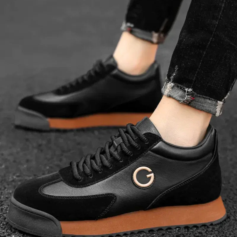 A person wearing sleek sneakers with brown soles, a metallic G logo, and luxe leather suede accents stands on a textured black surface, dressed in black jeans with frayed hems—perfect for the urban professional.