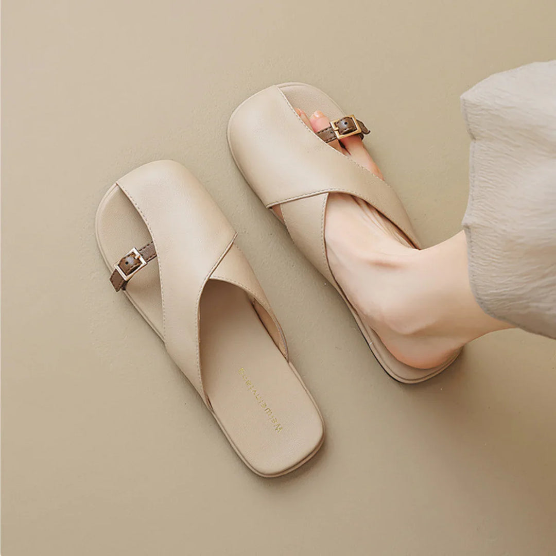 A person wearing chic slides with crisscross straps and a small buckle detail; one sandal is on the foot, the other on the floor. The neutral beige background makes these perfect additions to any summer wardrobe.