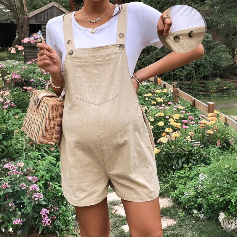 A woman in a white t-shirt and beige maternity overalls stands in a garden with colorful flowers. She carries a woven bag and wears layered necklaces. An inset shows a close-up of the button detail on the comfortable maternity overalls.