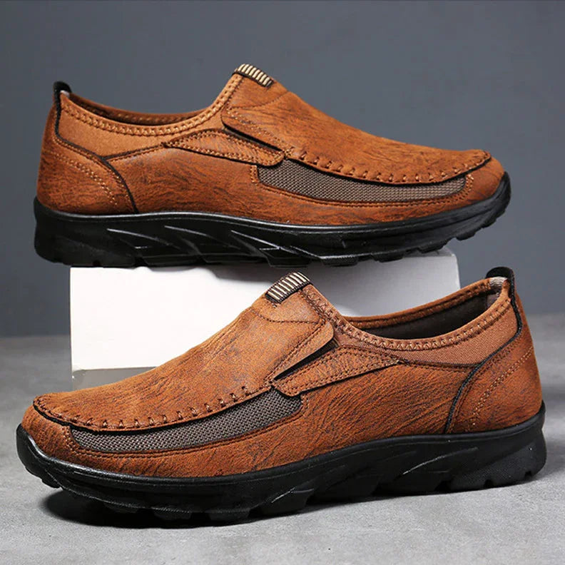 A pair of brown Men Leather Casual Shoes with black soles, displayed against a gray background. One shoe rests flat while the other is propped up, highlighting the comfortable fit and detailed stitching of these breathable loafers.