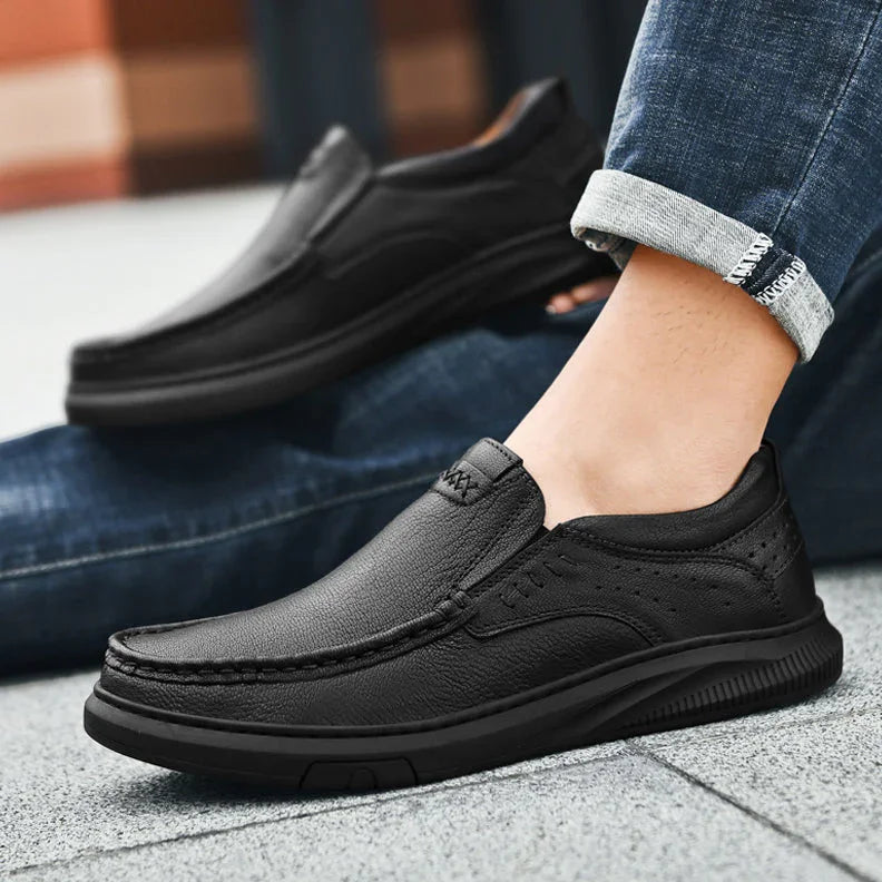 A person wearing dark blue jeans and men's cowhide leather slip-on shoes with a soft sole sits on the ground, one leg bent and the other stretched out. The shoes feature visible stitching and textured soles.