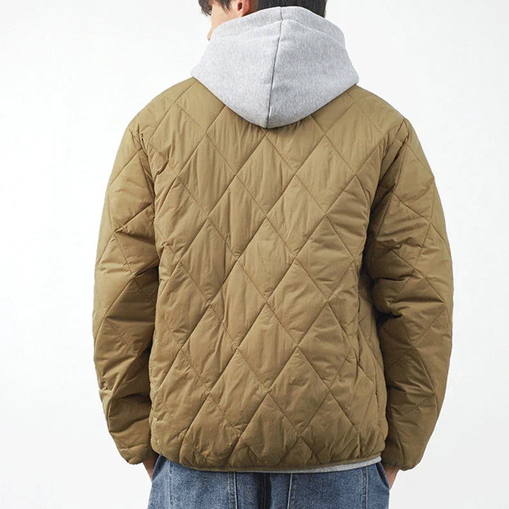 A person wearing a tan, lightweight insulating, diamond-stitched jacket over a gray hooded sweatshirt, seen from behind. The individual has short hair and is also wearing blue jeans. The background is plain and white.