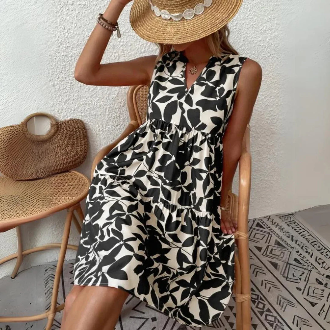 A woman in a sleeveless black and white leaf-print dress, made from lightweight breathable fabric, sits on a chair holding a straw hat. She accessorizes with bracelets and a necklace, surrounded by a woven bag and patterned rug—perfect for your summer wardrobe.