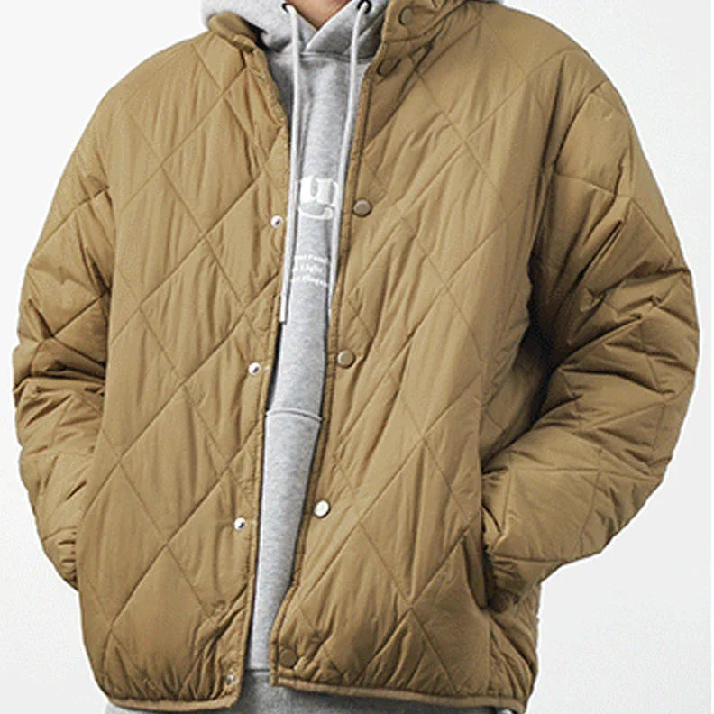 A person wearing a light brown, diamond-stitched pattern, quilted jacket over a grey hooded sweatshirt stands against a plain white background. Their hands are in the padded jacket pockets.
