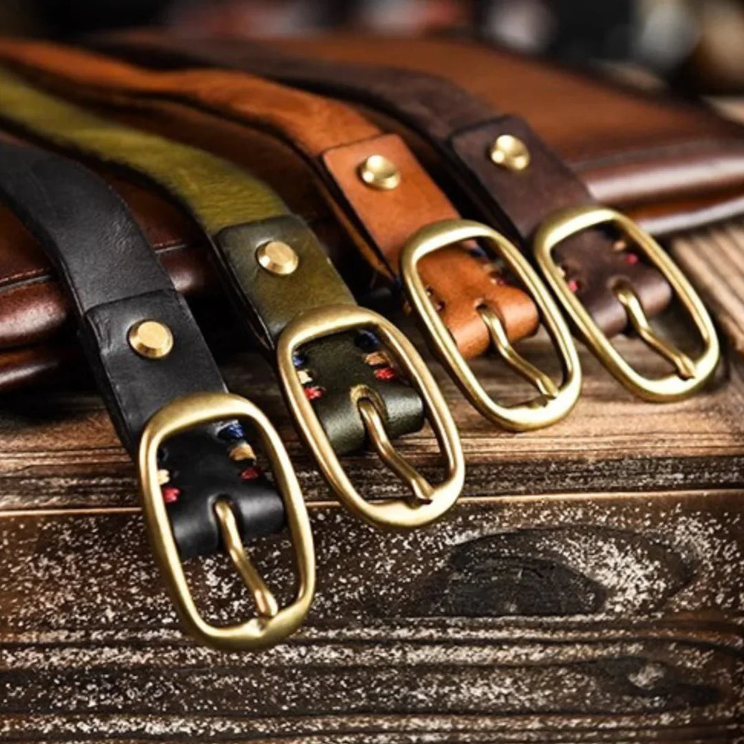 Four leather belts in black, green, brown, and dark brown with gold buckles are laid side by side on a wooden surface, showcasing country fashion style. The belts feature visible stitching near the buckles and metal rivets.