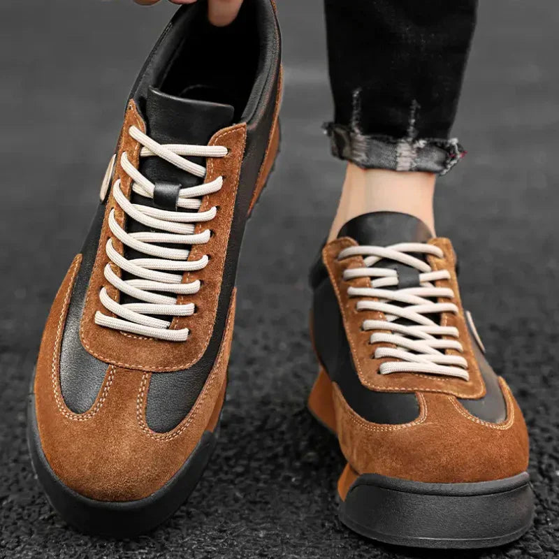 A person wearing black pants displays a pair of sleek sneakers crafted from luxe leather suede with white laces while standing on a dark textured surface.