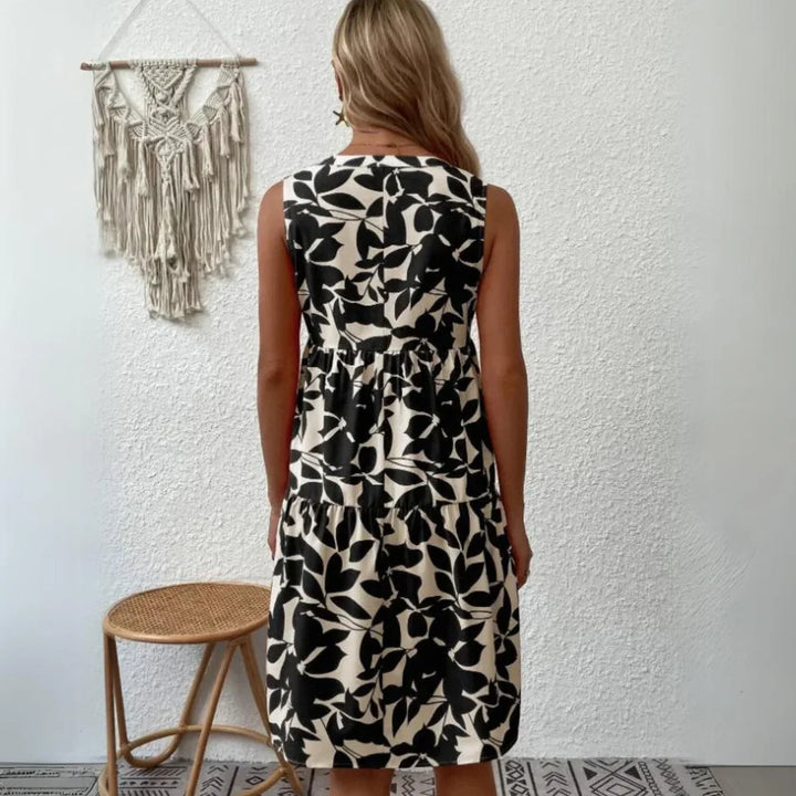 A woman with long blonde hair stands facing a white wall, wearing a sleeveless dress in a black and white leaf pattern—perfect for any summer wardrobe. A woven chair and macramé wall hanging add texture on the left side.