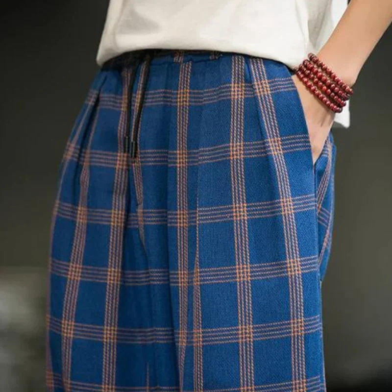 Person wearing blue plaid pants with orange lines—a bold fashion statement—paired with a white shirt and a red beaded bracelet, one hand in their pocket, shown from the waist down.