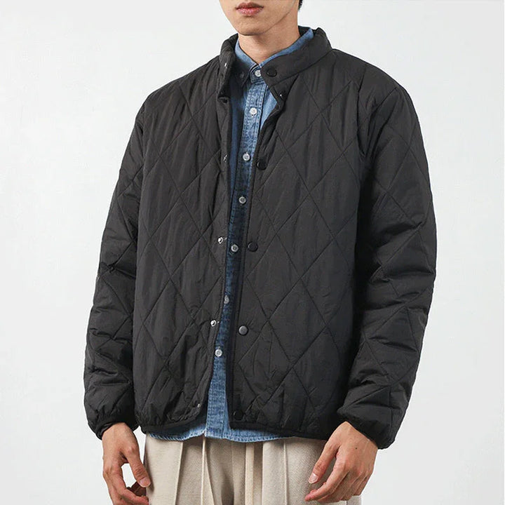 A person wearing a black, diamond-stitched padded jacket over a blue button-up shirt and beige pants stands against a plain white background. The jacket has a high collar and snap buttons. The persons face is partially out of frame.