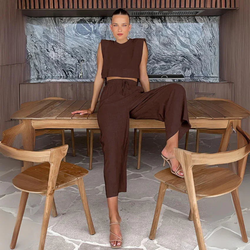 A woman in a sleeveless brown cropped top and matching wide-leg pants sits on a wooden table, one leg up, surrounded by wooden chairs in a modern room with a marble backsplash and Belgian Linen Blend accents.