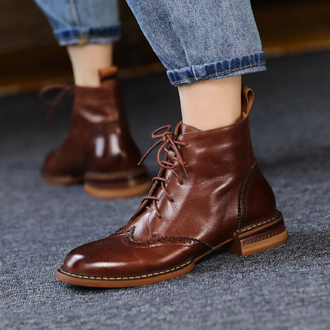 A person wearing rolled-up blue jeans and brown genuine leather lace-up ankle boots stands on a carpeted floor. The boots, with decorative stitching and a small heel, make a classic statement for the urban trendsetter.