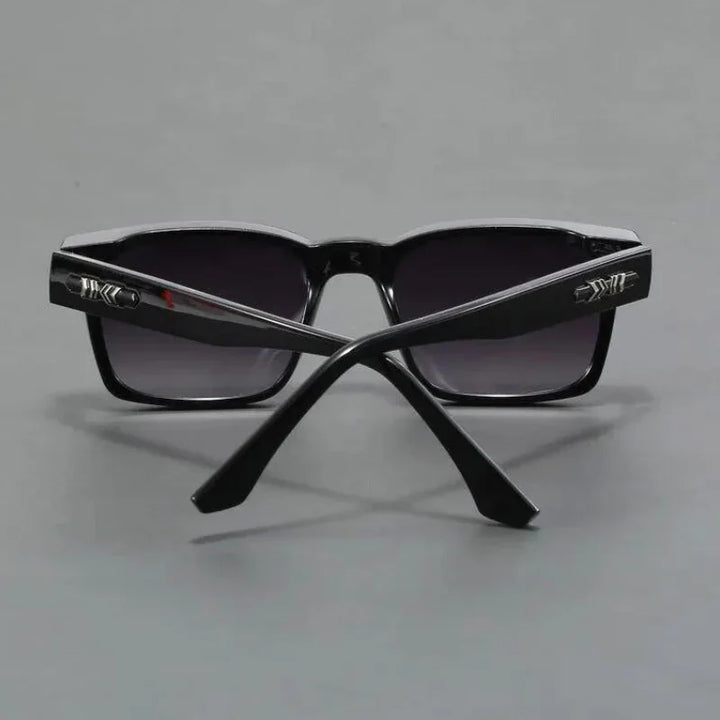 A pair of black sunglasses with dark lenses, shown from the back with the arms crossed. The durable frames feature metallic decorative details near the hinges. The background is a plain gray surface.