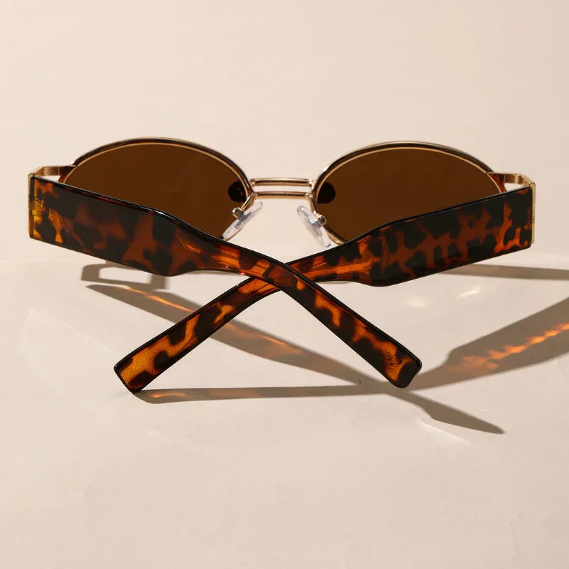 A pair of brown retro oval sunglasses with a durable metal frame and thick, tortoiseshell-patterned arms crossed at the ends, displayed on a light beige surface with soft shadows.