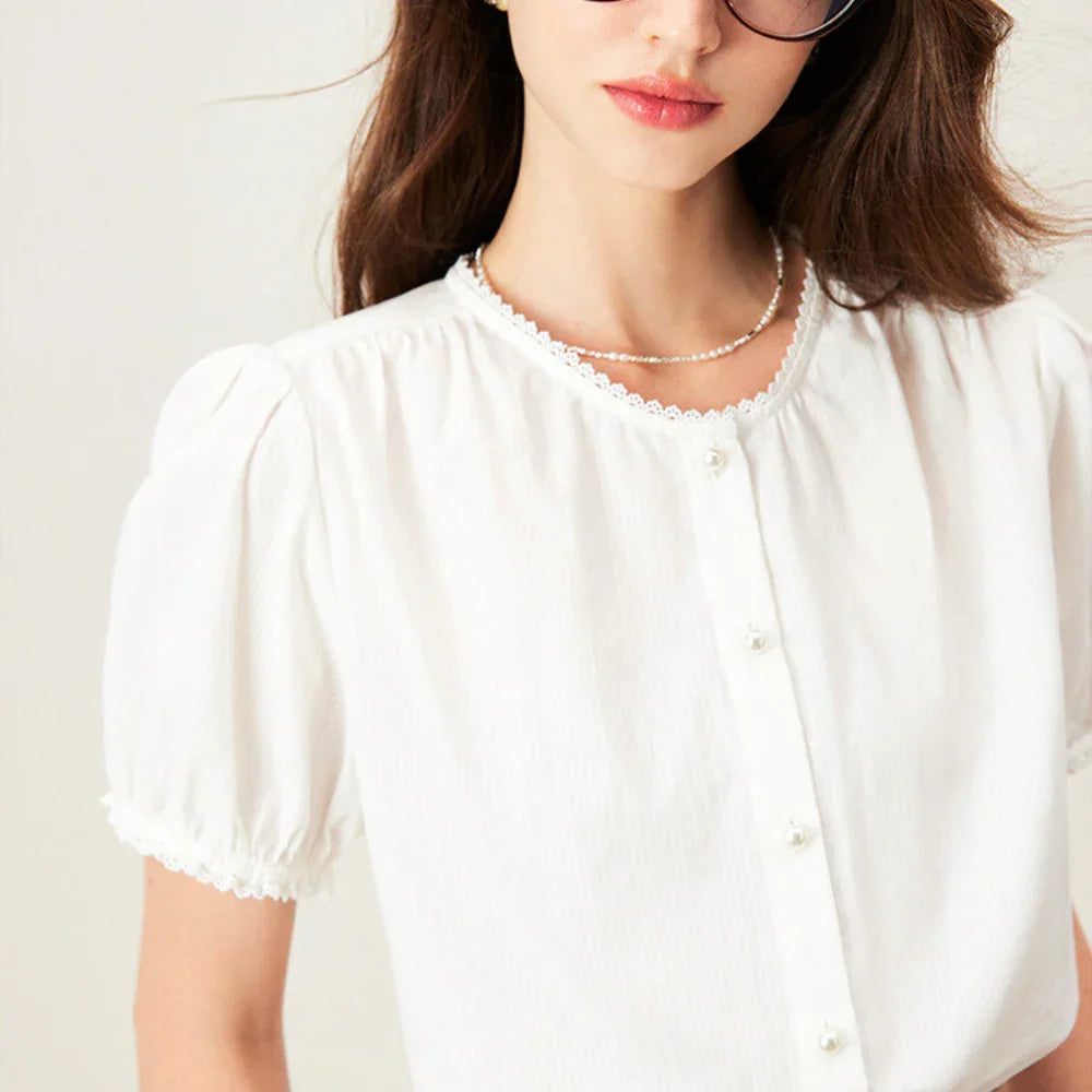 A woman with long brown hair wears sunglasses and a white lace trim blouse with puffed short sleeves and pearl buttons. She completes her look with a delicate pearl necklace. Only her upper body is visible.