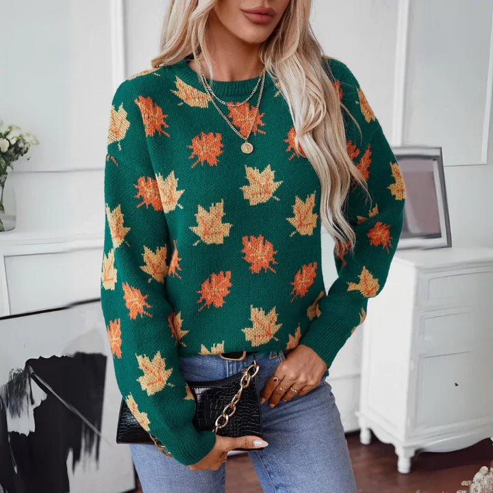 A woman with long blonde hair wears a comfortable green sweater with a maple leaf pattern in orange and yellow, light blue jeans, and holds a black purse with a gold chain. She stands indoors near white furniture and framed art.