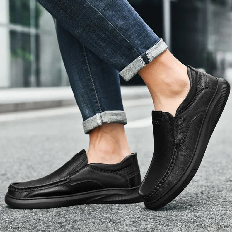 A person in rolled-up blue jeans stands on a street, showcasing Men's Cowhide Leather Slip-On Shoes with a soft, textured sole. The focus is on the footwear and lower legs.