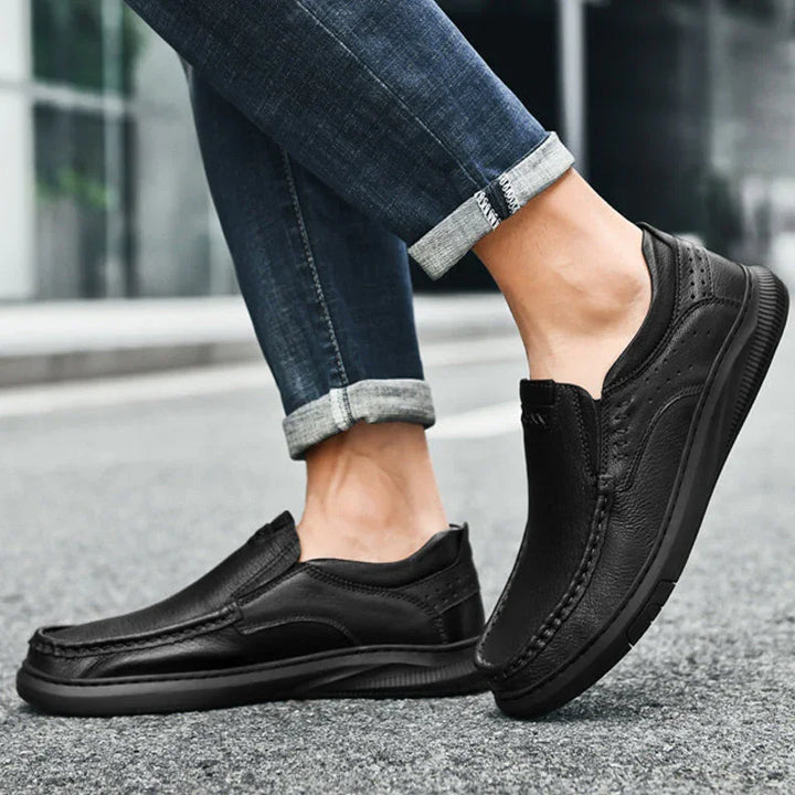 A person in rolled-up blue jeans stands on a street, showcasing Men's Cowhide Leather Slip-On Shoes with a soft, textured sole. The focus is on the footwear and lower legs.