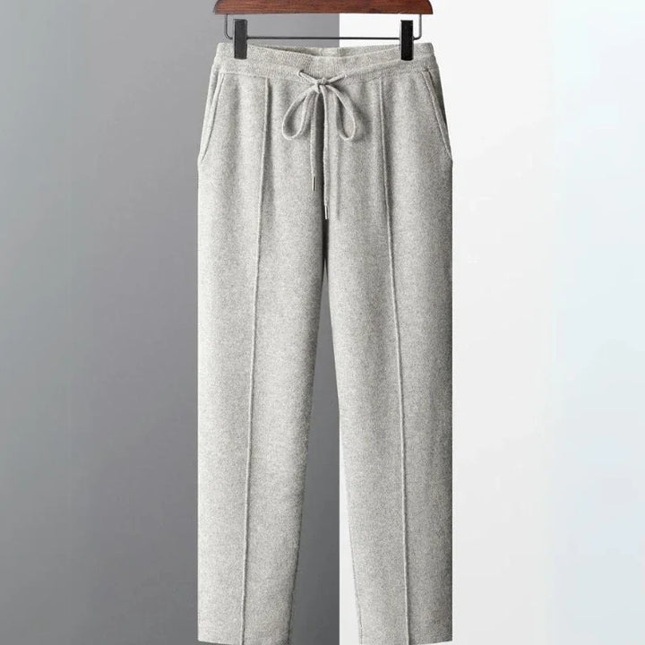 Light gray, soft-textured comfortable pants with an elastic waistband, front tie drawstring, and side pockets, hanging on a wooden hanger against a two-tone gray and white background.