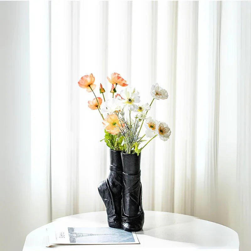 A black ballet-inspired ceramic vase shaped like pointe shoes holds orange and white flowers on a round white table, with a folded newspaper nearby and sheer white curtains in the background—perfect for contemporary chic décor.