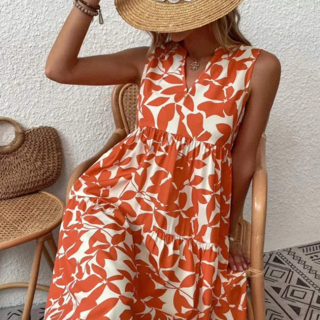 A woman sits on a chair in a sleeveless, orange and white leaf-patterned dress made from lightweight breathable fabric. She holds a wide-brimmed straw hat over her face and accessorizes with bracelets, a necklace, and a woven bag.