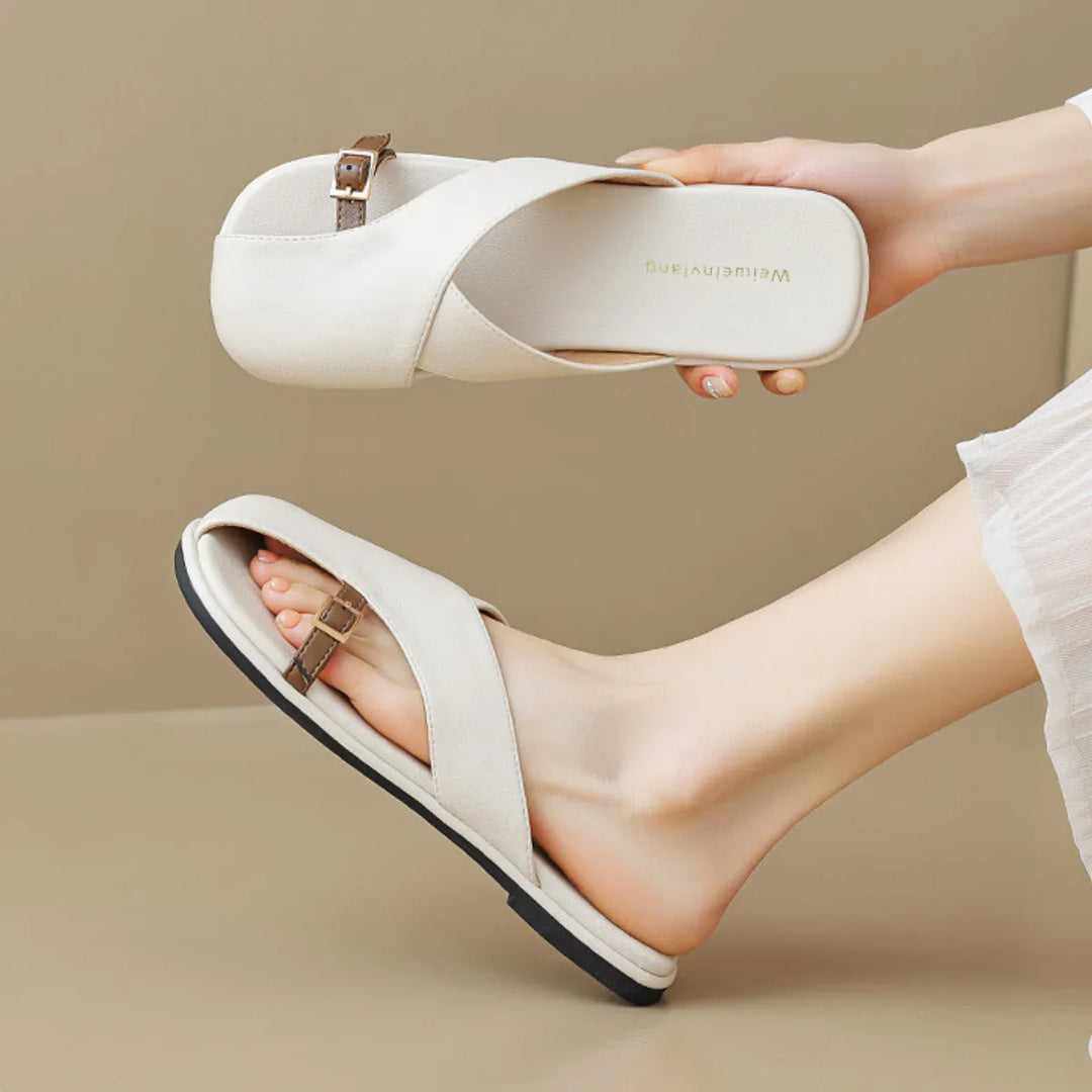 A person wearing chic slides—white open-toe slip-ons with a small buckle detail, holding one sandal in their hand. The beige background and light-colored outfit make these sandals perfect for any summer wardrobe.