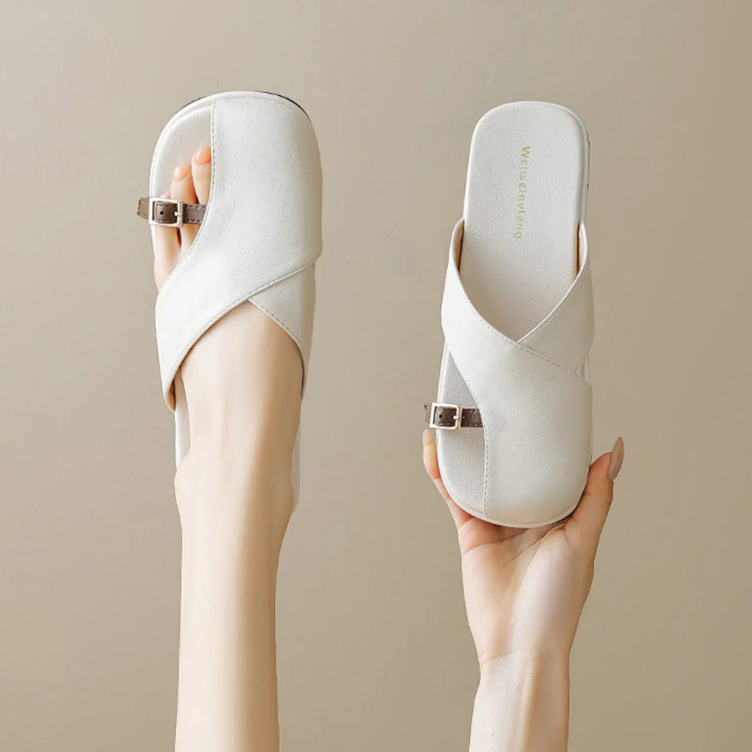 A pair of chic slides with white crisscross straps and small side buckles, held up by two hands against a neutral beige background—the perfect addition to your summer wardrobe.