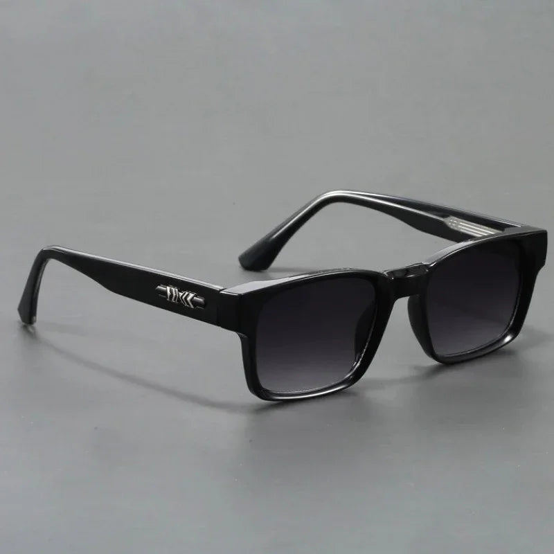 A pair of black rectangular sunglasses with modern design, featuring dark lenses and a silver decorative detail on the arms, displayed on a plain gray surface.