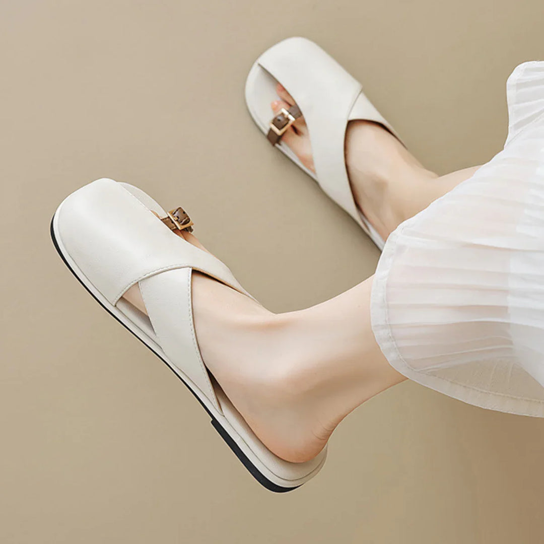 A person wearing chic white leather slides with crisscross straps and small buckles, paired with a white pleated skirt, against a beige background—perfect for a summer wardrobe.