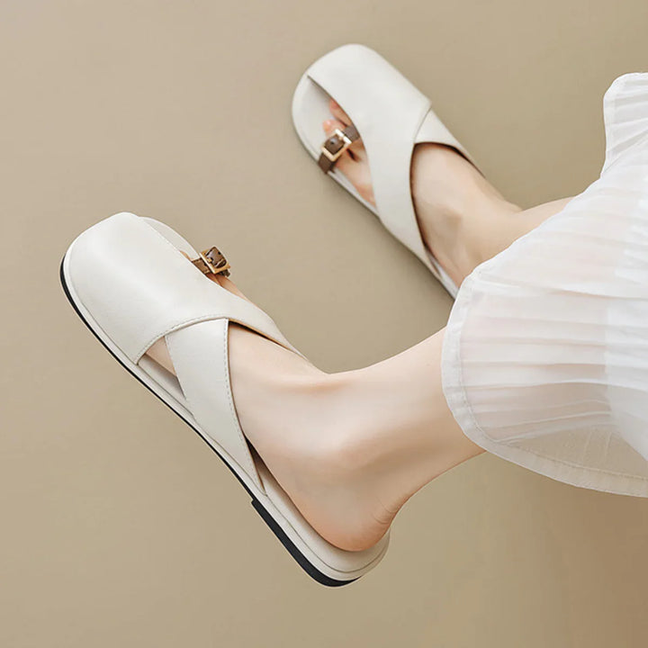 A person wearing chic white leather slides with crisscross straps and small buckles, paired with a white pleated skirt, against a beige background—perfect for a summer wardrobe.