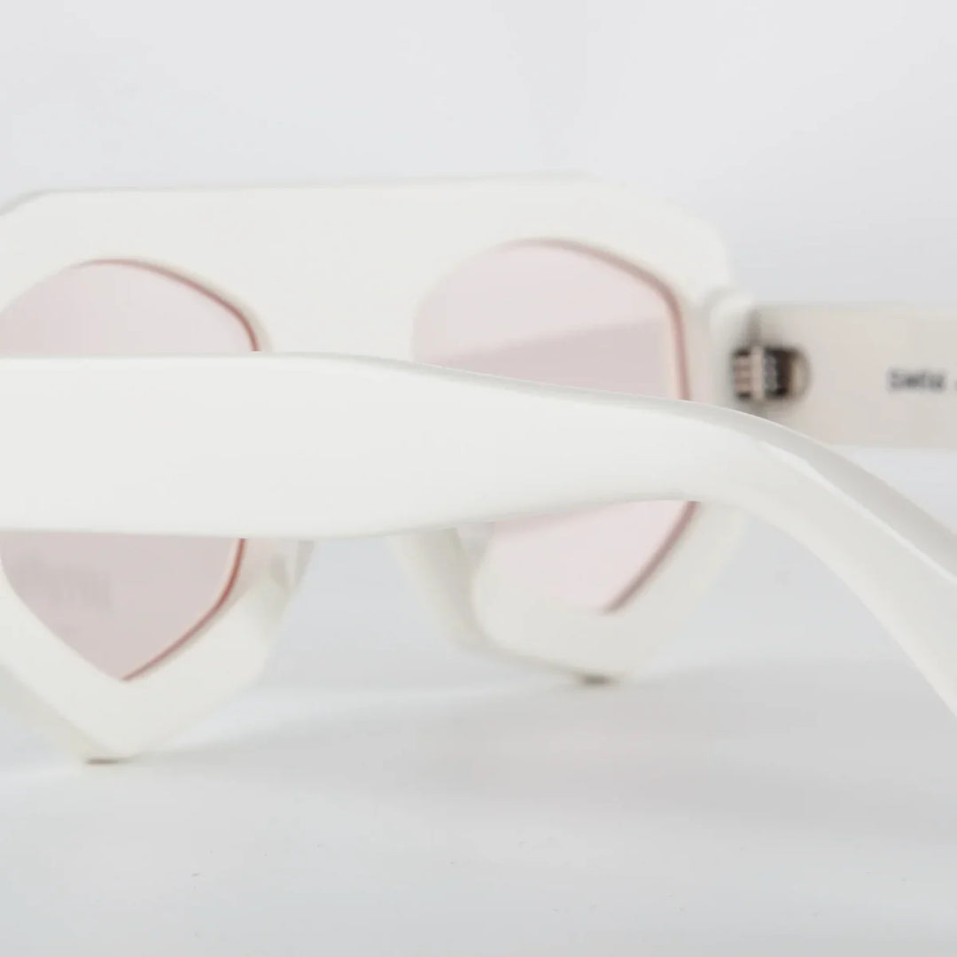 Close-up of white acetate sunglasses with pink-tinted lenses and a geometric frame, focusing on the temple and frame against a plain white background.