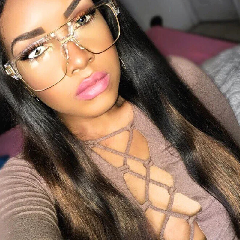 A woman with long straight hair wears large geometric sunglasses with UV400 protection and a brown top with a crisscross lace-up front. She has bold eyebrows and pink lipstick, posing indoors on a bed with pink covers.