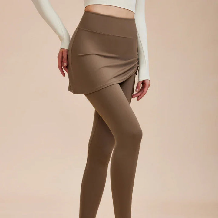 A person wearing high-waisted leggings with a matching beige skirt and a long-sleeved, fitted white crop top stands against a neutral background. Only the lower torso and legs are visible—perfect for an autumn wardrobe update.