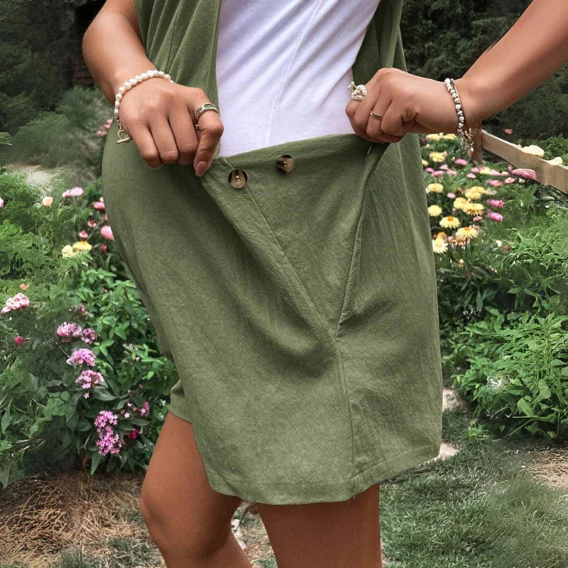 A person in a green maternity overall dress and white top stands in a garden, pulling at the side to show button details. Colorful flowers and greenery are visible in the background. The persons face is not shown.