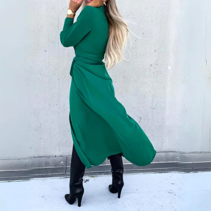 Woman with long blonde hair wears a flowing long-sleeve wrap dress and black heeled boots, standing on a light-colored surface turned away from the camera against a plain, light gray background—a touch of high fashion elegance.
