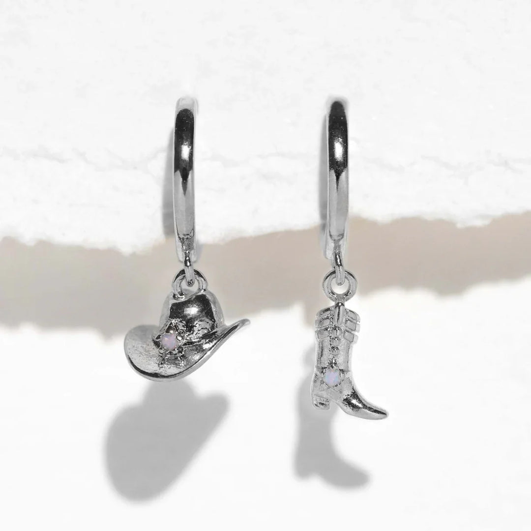 Two silver hoop statement earrings hang against a white textured background; one earring features a cowboy hat charm and the other a cowboy boot charm, each adorned with a small clear gemstone.