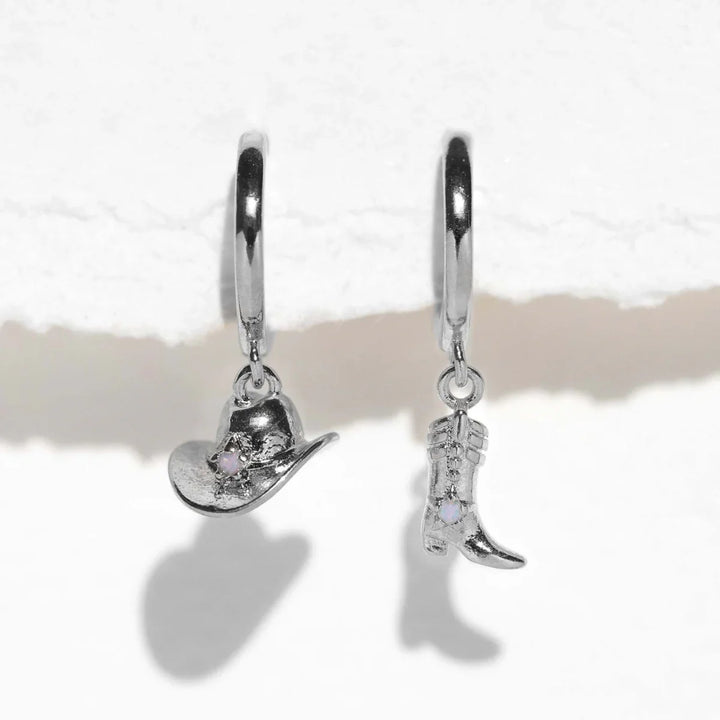 Two silver hoop statement earrings hang against a white textured background; one earring features a cowboy hat charm and the other a cowboy boot charm, each adorned with a small clear gemstone.