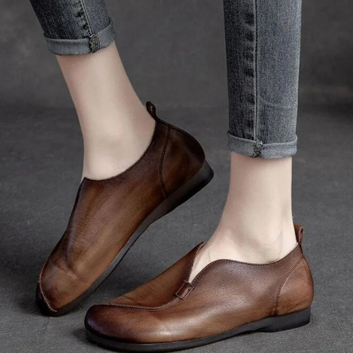 A person wearing rolled-up jeans and brown leather slip-on shoes with a cushioned insole, standing on a gray surface. The shoes have a minimalist design, supple leather, and a slight sheen with a low heel.