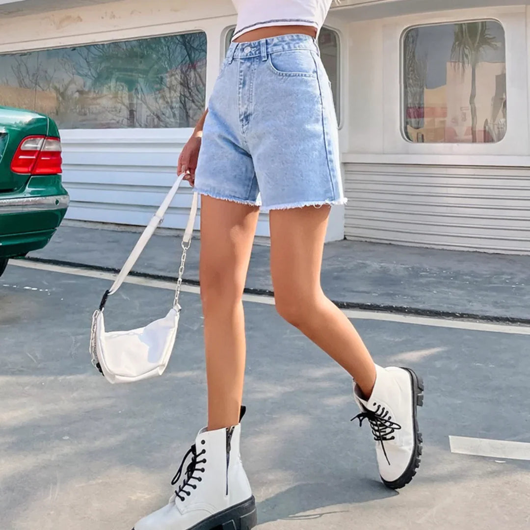 A person wearing a white crop top, high-waist denim shorts, white combat boots, and carrying a white handbag is walking outdoors near a green car and a building with large windows.
