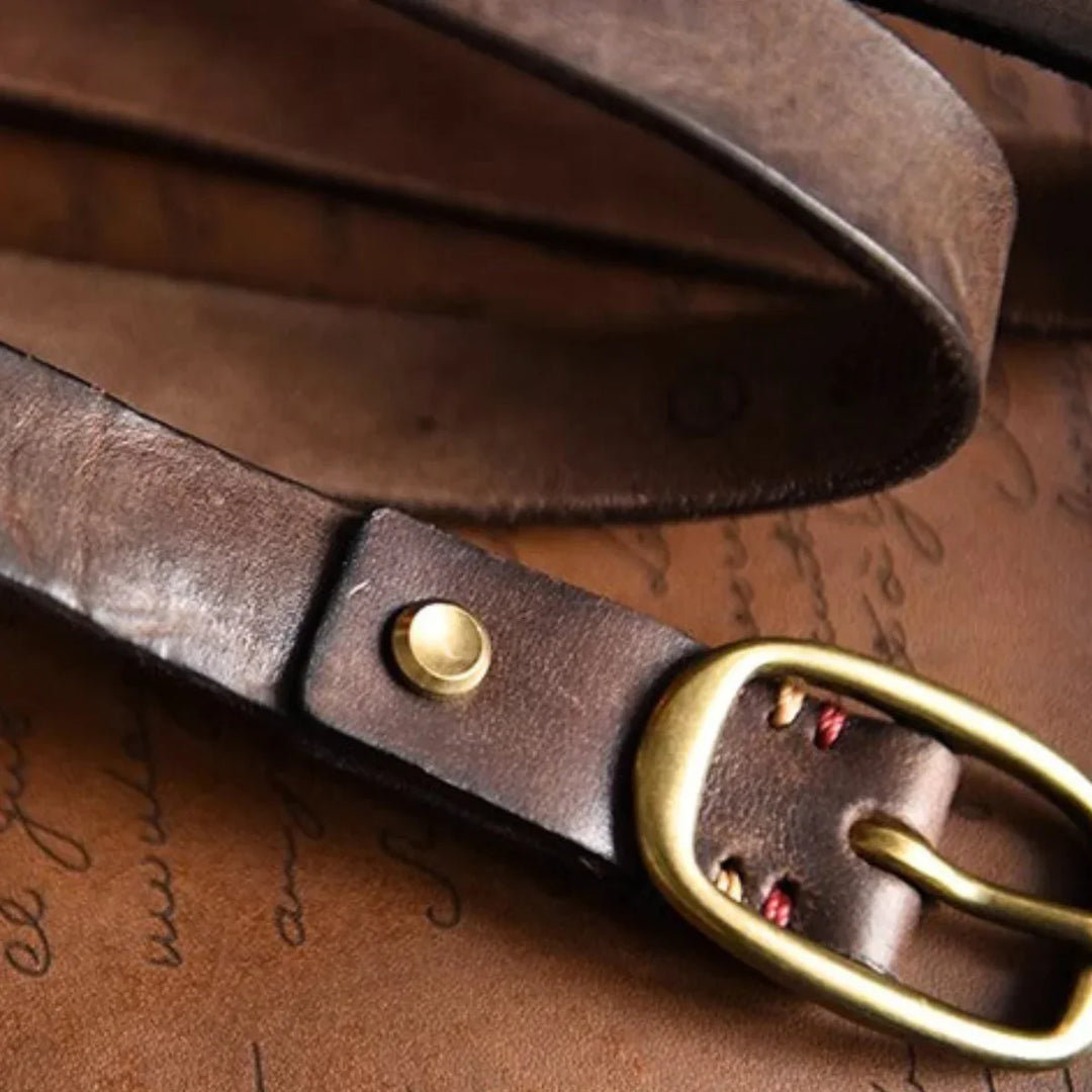 Close-up of a brown leather belt with a brass buckle, resting on a textured leather surface with faint cursive writing in the background—a timeless piece for country fashion enthusiasts.