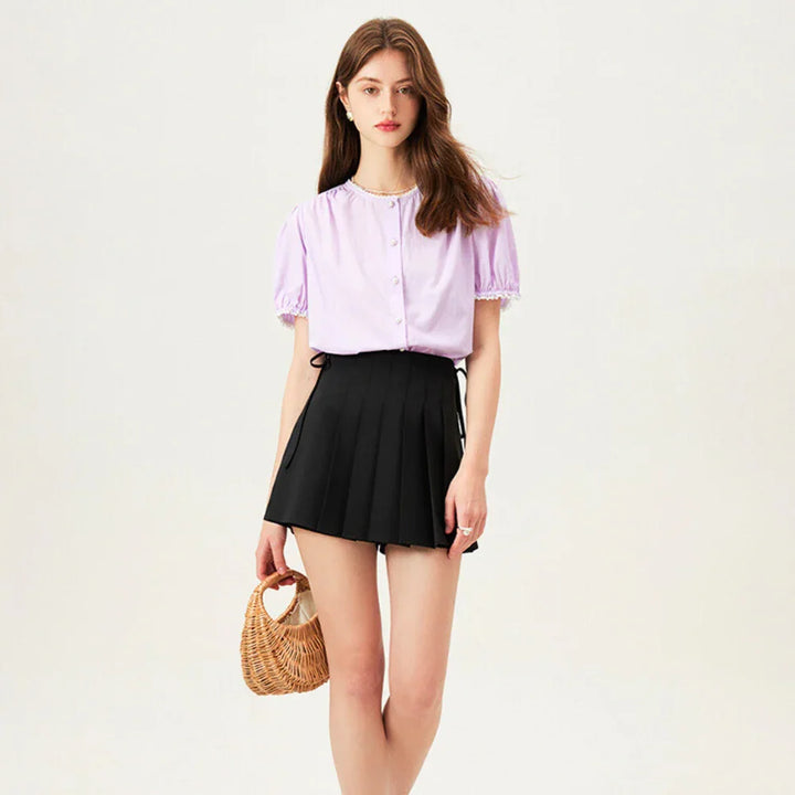 A young woman stands against a plain background, wearing a light purple button-up blouse with short puff sleeves and a black pleated mini skirt. She holds a small woven handbag and has long, loose hair.
