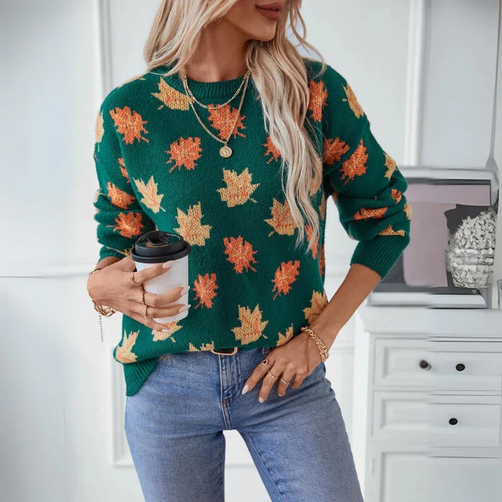 Woman wearing a comfortable green sweater with a soft knit material and subtle maple leaf pattern, blue jeans, and layered necklaces, holding a to-go coffee cup in a bright room with white furniture in the background.