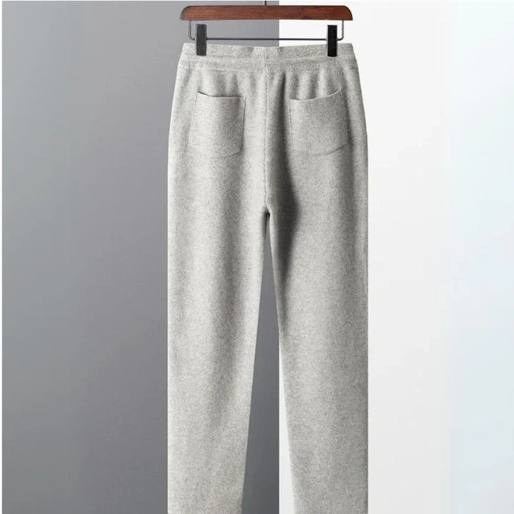 A pair of light gray, comfortable pants with two back pockets and durable construction hangs on a wooden hanger against a split gray and white background.