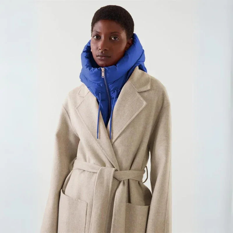 A person wearing a beige belted coat over a bright blue puffer jacket with a hooded design, standing against a plain light background.