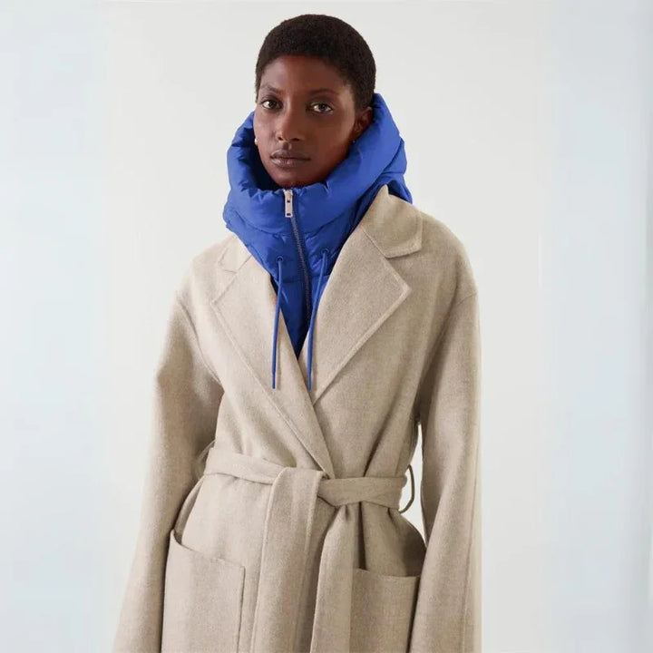 A person wearing a beige belted coat over a bright blue puffer jacket with a hooded design, standing against a plain light background.
