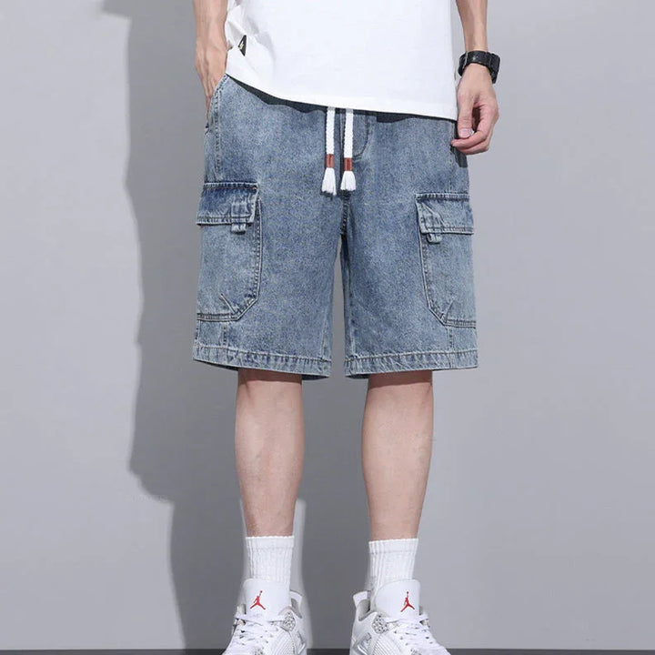 A person wearing light blue mens denim shorts with an elastic waist, a white T-shirt, white socks, and white sneakers stands against a gray background. Only the lower half of their body is visible.