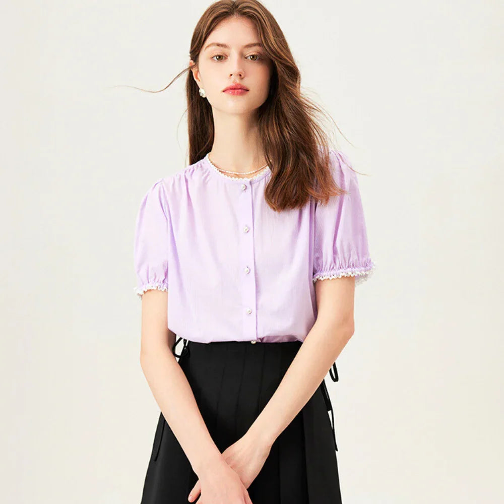 A young woman with long brown hair wears a light purple lace trim blouse with pearl-like buttons and a black high-waisted skirt, standing against a plain light background.